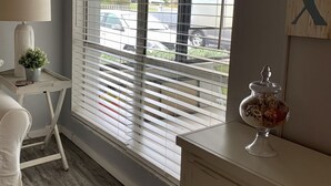 Una habitación con un ventanal con persianas, una mesita con una lámpara, un frasco decorativo y una silla.