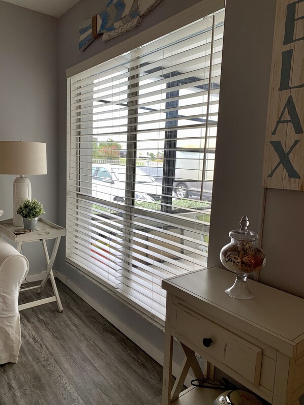 A room with a large window with blinds, a small table with a lamp, a decorative jar, and a chair.