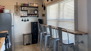 A compact kitchen with a refrigerator, a sink, and a countertop. There are bar stools and a window with blinds.