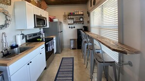 A compact kitchen with white cabinets, a stainless steel refrigerator, a microwave, and a wooden countertop. There are bar stools and a window with blinds.