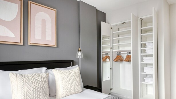 A neatly arranged bedroom with a bed, pillows, a bedside lamp, and an open walk-in closet with hanging clothes. - Modern Waterloo 2BR open plan with Balcony on Lower Marsh, by Blueground (London)