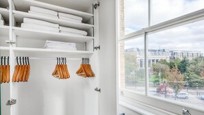 A white closet with shelves for folded towels and wooden hangers for clothes. A window reveals a cityscape with buildings and trees. - Modern Waterloo 2BR open plan with Balcony on Lower Marsh, by Blueground (London)
