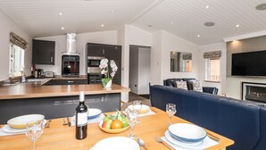 A modern kitchen with a central island, a dining area with a table set for four, and a living area with a sofa and TV. - Ridgway Lodge (Windermere)