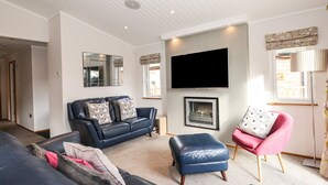 A living room with a fireplace, a flat-screen TV, a blue leather sofa, a pink armchair, and a large mirror. - Ridgway Lodge (Windermere)