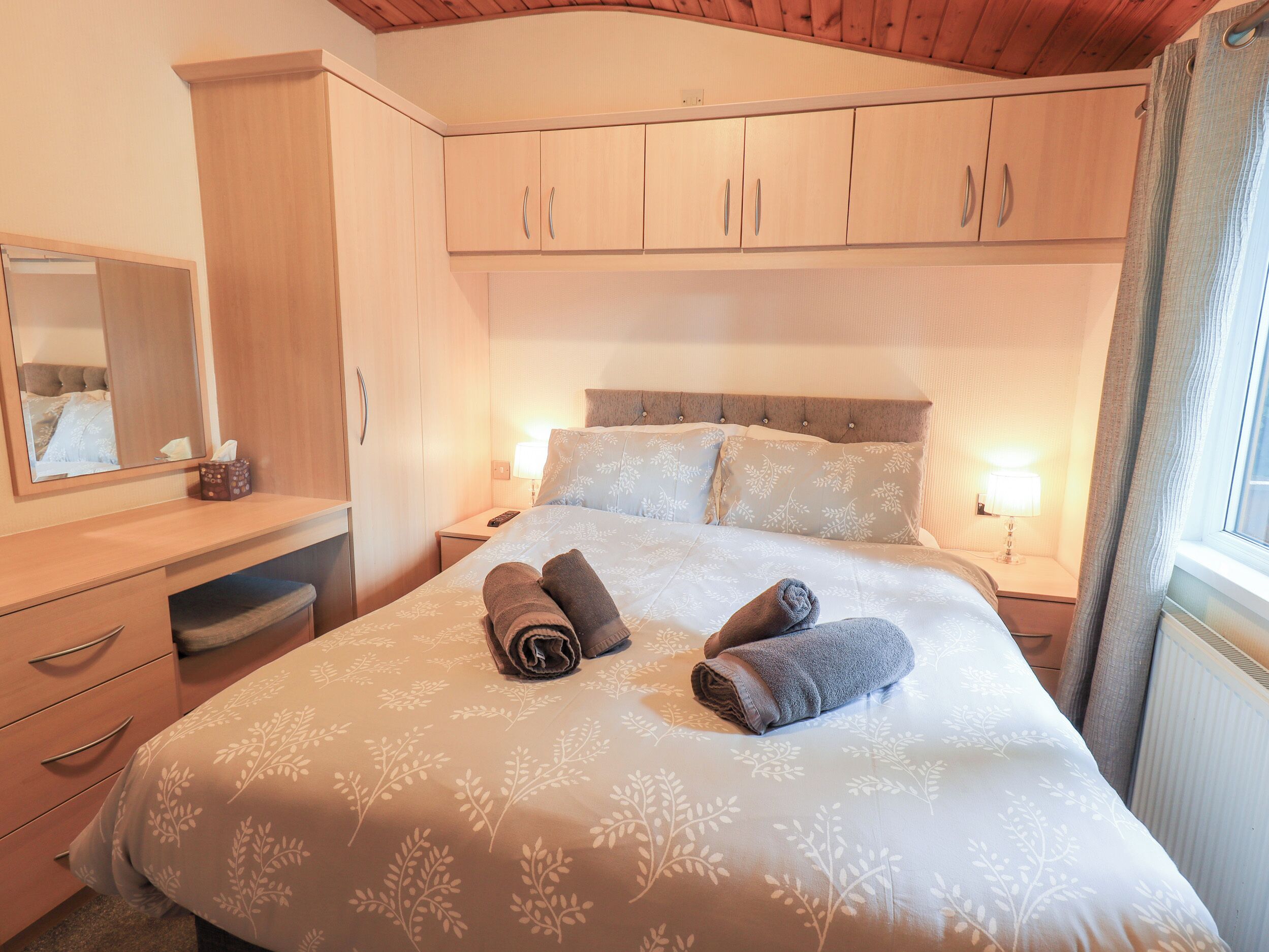 A neatly made bed with two rolled towels, a bedside table with a lamp, and a mirror on the wall.