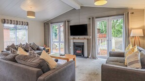 Ein Wohnzimmer mit Kamin, einer Sofagarnitur, einem Fernseher und großen Fenstern mit Vorhängen.