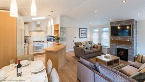 A modern living room with a fireplace, a dining area with a table set for two, and a kitchen with white cabinets and a central island. - Broad Larch Lodge (Windermere)