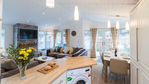 A modern living room with a sofa, a dining area with a table set for two, a washing machine, and a TV mounted on the wall. - Broad Larch Lodge (Windermere)