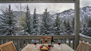 Et træbord dækket med vinglas, en flaske og et ostebord på en balkon med udsigt til en sneklædt skov.