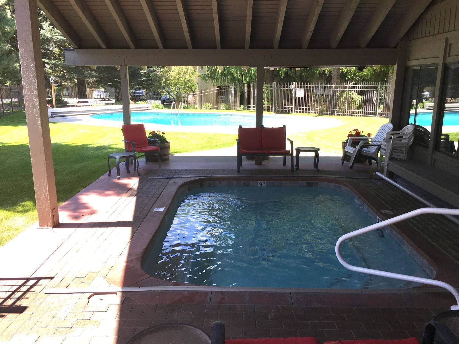 A covered outdoor swimming pool area with a hot tub, lounge chairs, and a view of a larger pool in the background.