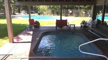 A covered outdoor swimming pool area with a hot tub, lounge chairs, and a view of a larger pool in the background.