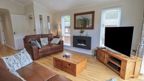 A living room with a leather sofa, a wooden coffee table, a flat-screen TV on a wooden stand, and a fireplace. - Wansfell Retreat Lodge (Windermere)
