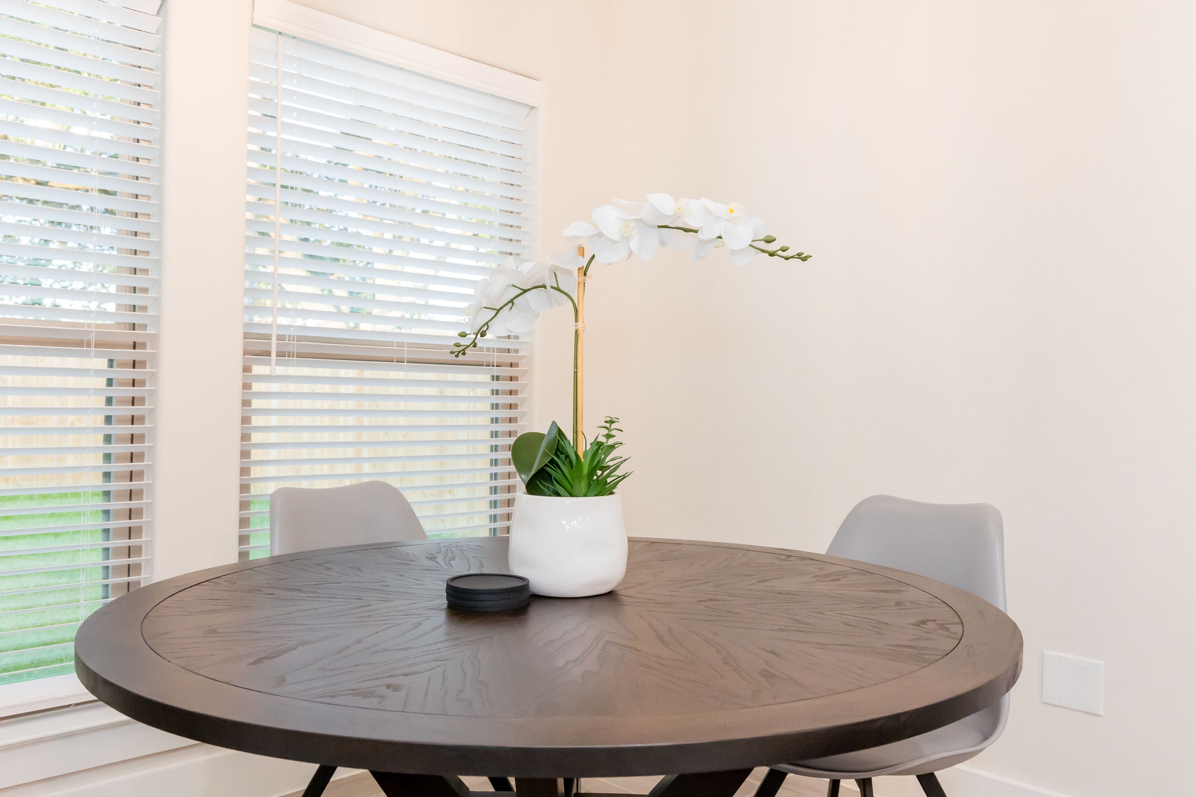 A round dining table with a white potted plant and a black object on it, surrounded by two chairs, in a room with large windows and blinds.