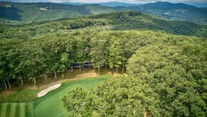Luchtbeeld van een golfbaan, omgeven door dicht bos en bergen.
