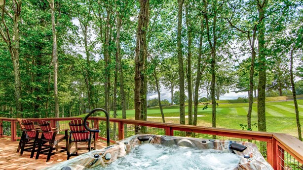 Een hottub op een houten terras met stoelen, omringd door een bos en een golfterrein op de achtergrond.