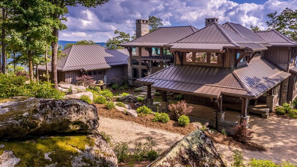 Un grand bâtiment de style rustique, doté de plusieurs toits et d’une cheminée en pierre, entouré d’un aménagement paysager naturel et de gros rochers.