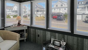 A cozy room with a wicker sofa, a small table with a lamp, and a view of a street through large windows with blinds.