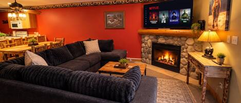 Una sala de estar acogedora con un sofá modular, una chimenea, un televisor mostrando series de Netflix y un comedor al fondo.