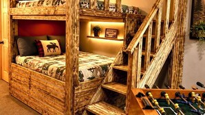 Una cama litera con escalera de madera, un pequeño sofá y una mesa de fútbol en una habitación con pared roja y luces colgantes.
