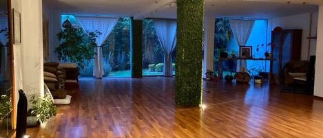 A spacious living room with wooden flooring, a central pillar, and large windows with curtains.