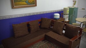 A corner sofa with brown cushions, a blue and white patterned wall, a small table with a green cabinet, and a chair with a yellow cushion. - Tempat Nginap 2BR Ciater Highland Resort (Ciater)