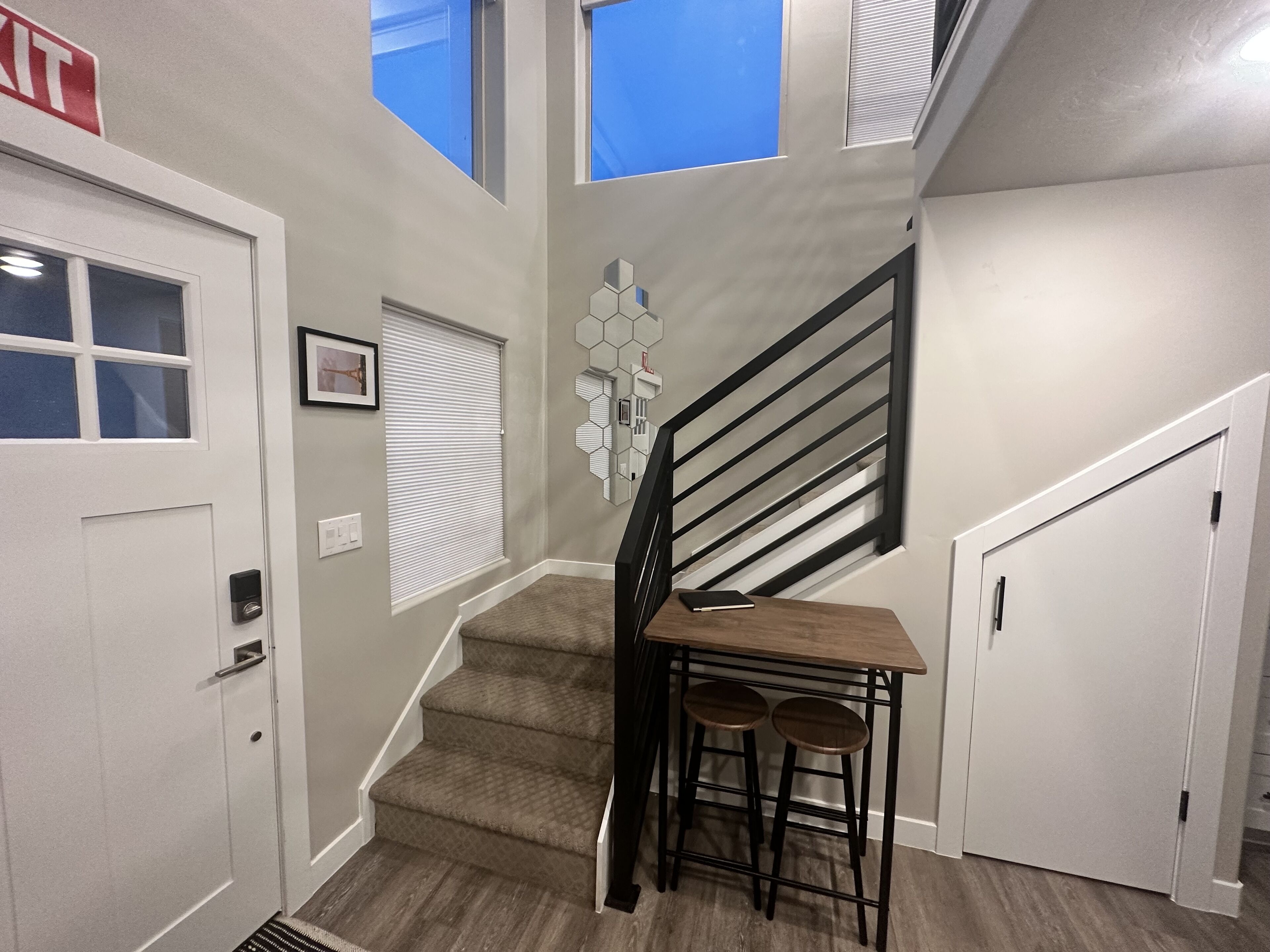 A staircase with a wooden table and two stools, a white door with a keypad, and a hexagonal wall decoration.