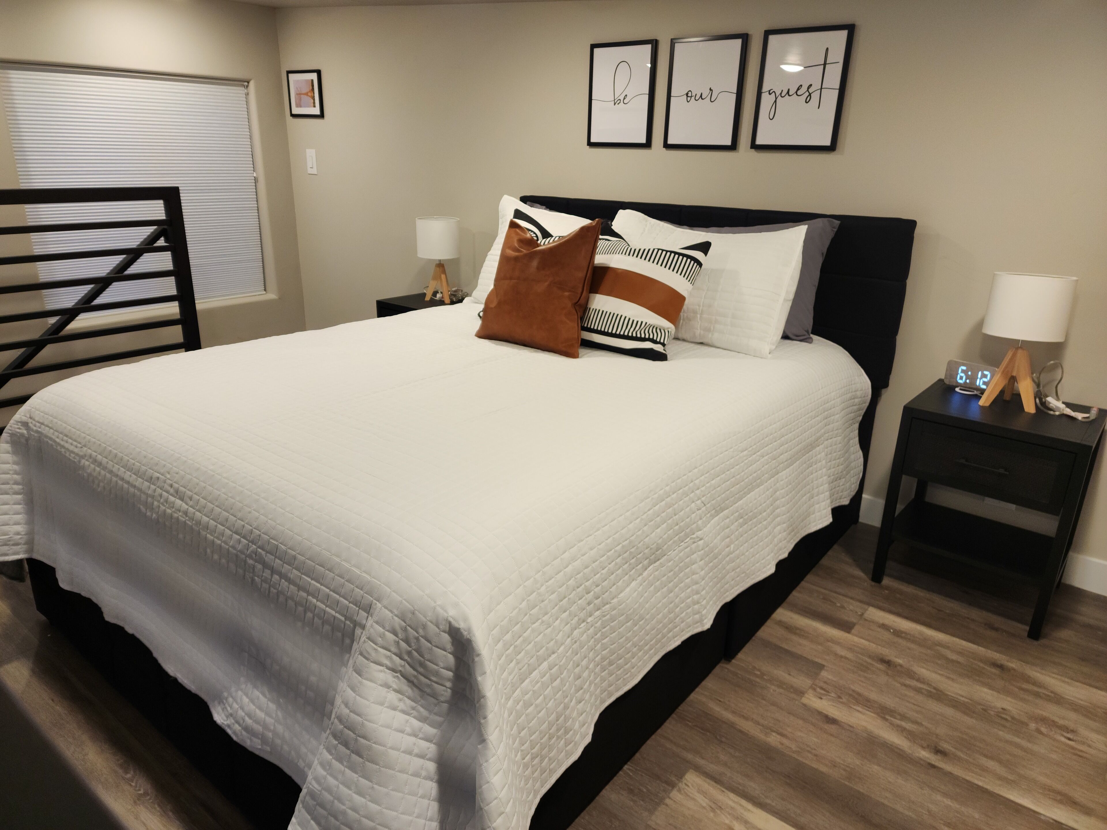 A neatly made bed with a white quilt, a wooden headboard, and a nightstand with a lamp and a clock.