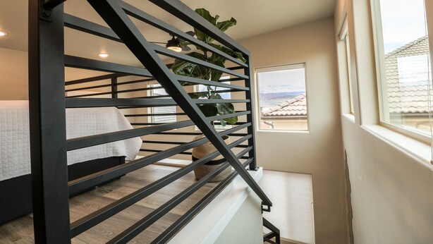 Una escalera con barandilla de metal negro, una cama con ropa de cama blanca, una planta en maceta y una ventana con vista a edificios.
