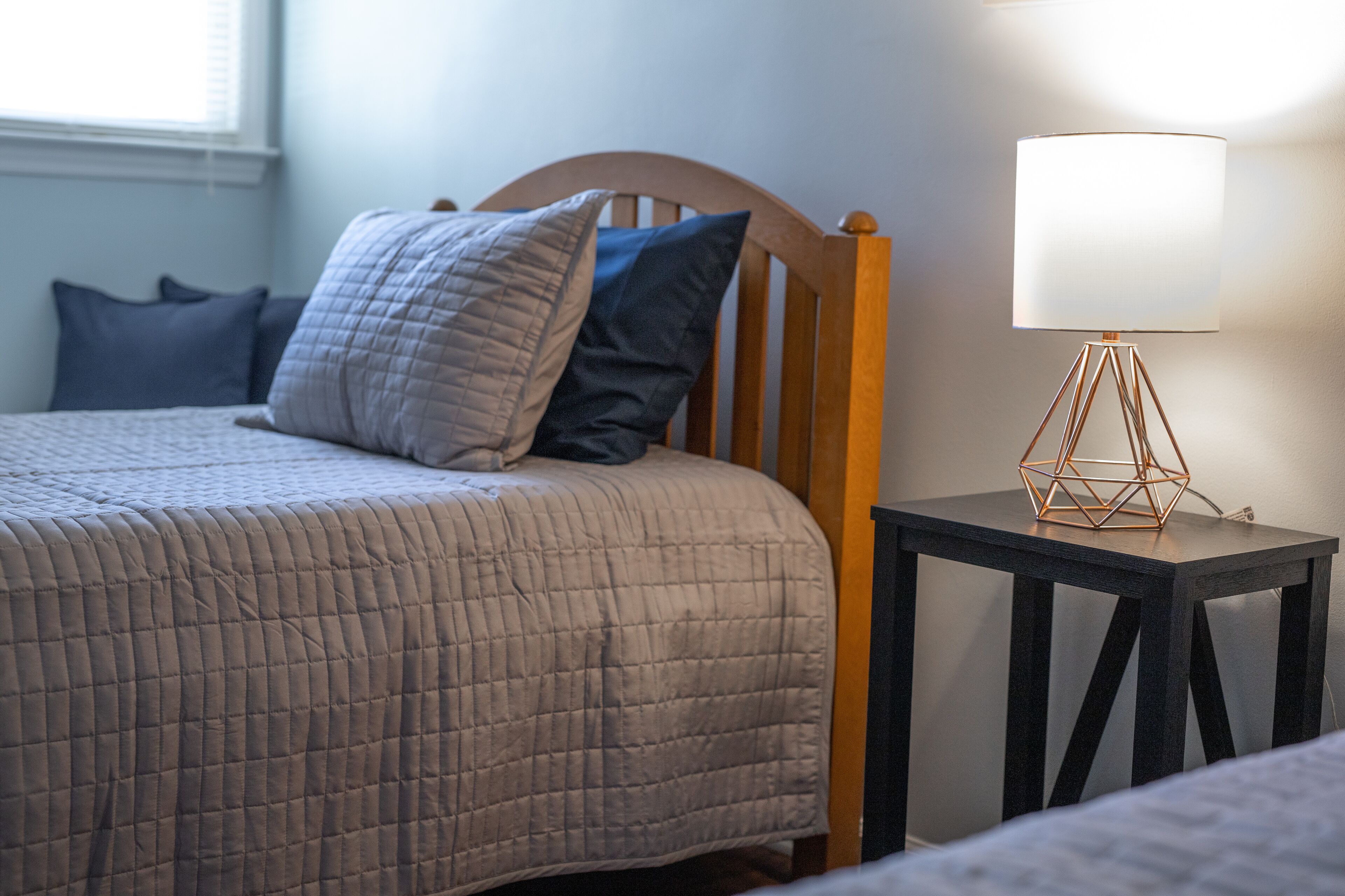 A neatly made bed with a quilted blanket and two pillows, a bedside table with a geometric lamp, and a window with blinds in the background.
