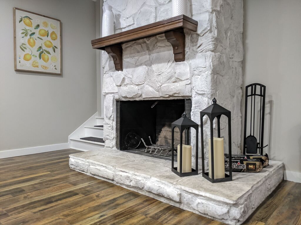 A stone fireplace with a wooden mantel, two black lanterns, and a framed picture of lemons on the wall.