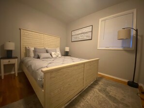 A bedroom with a wooden bed, two bedside tables, a floor lamp, a window with blinds, and a framed picture on the wall.