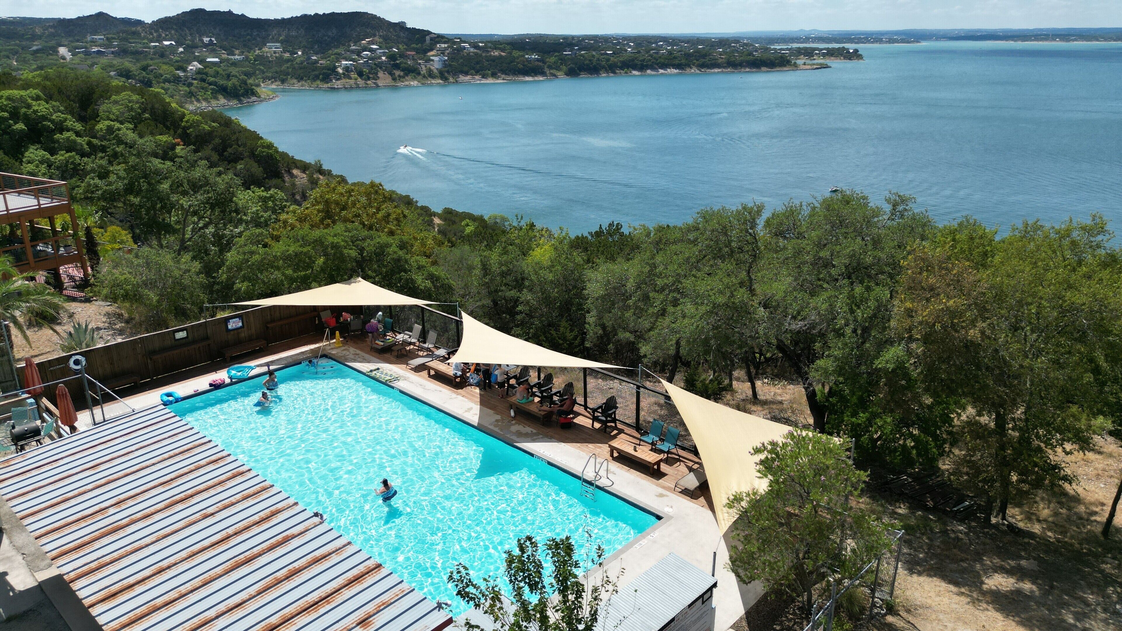 A pool with a wooden deck and lounge chairs, surrounded by trees and overlooking a body of water.