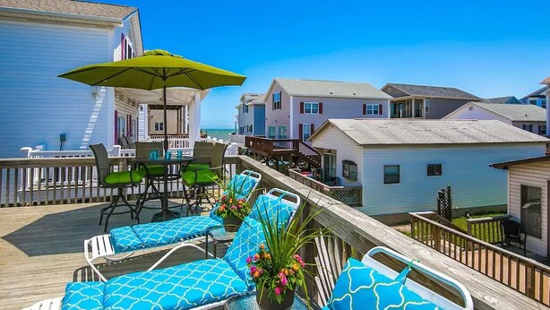 Um deck de madeira com cadeiras de descanso e uma mesa, com vista para uma área residencial com casas e um céu azul límpido.