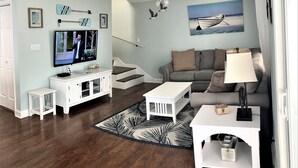 Uma sala de estar com televisão, móveis brancos, um sofá cinza, um tapete estampado e um ventilador de teto.