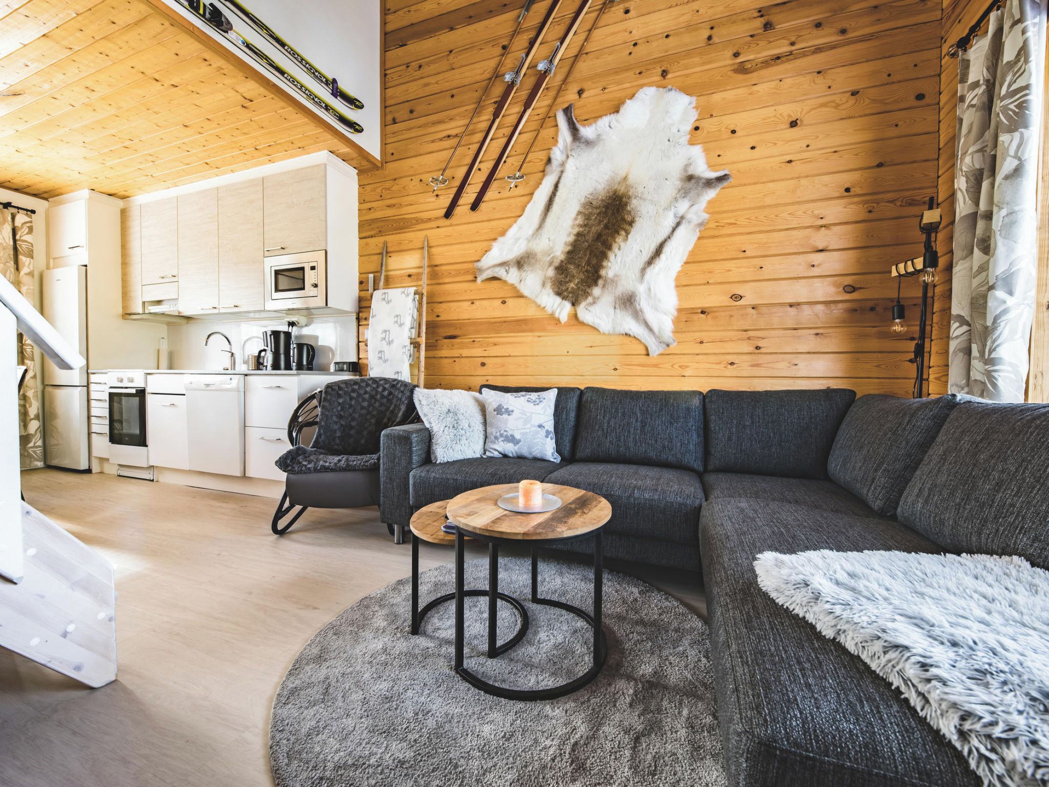 A modern living room with a wooden ceiling, a large fur rug, a grey sofa, a round coffee table, and a kitchen in the background.