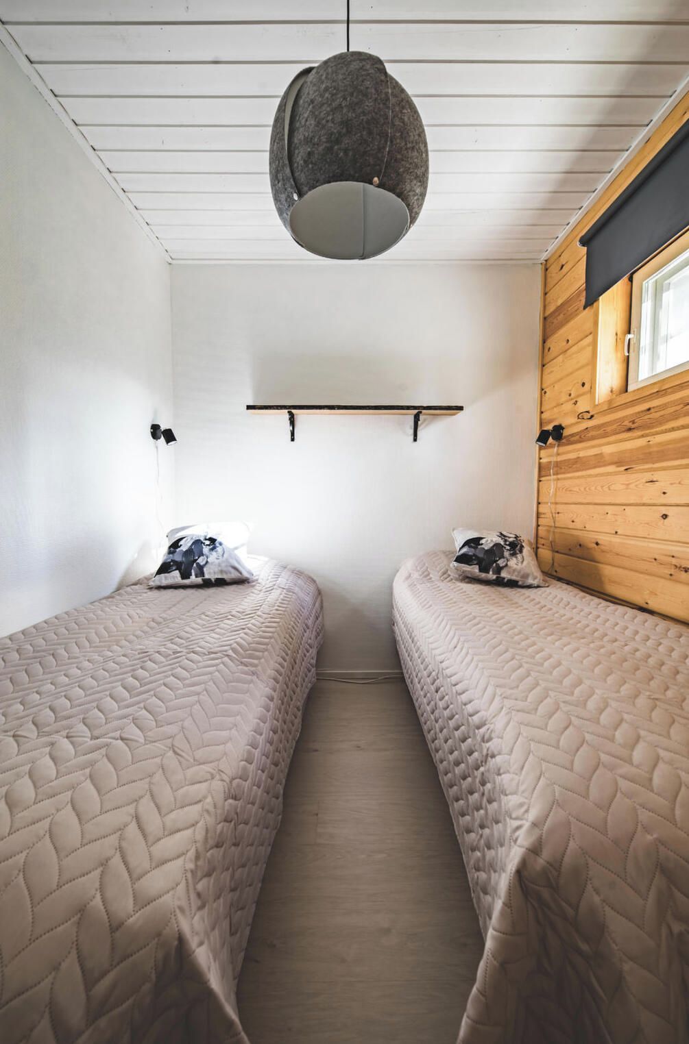 A room with two beds, a hanging light fixture, a wooden wall, and a window.