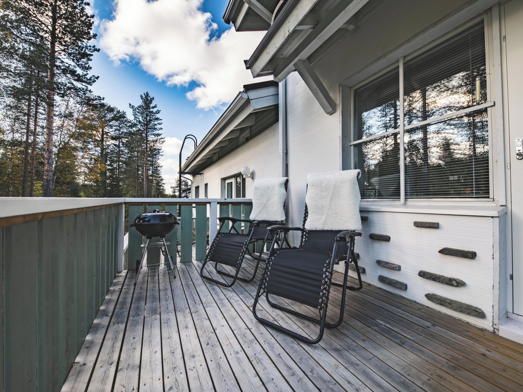 A wooden deck with a barbecue grill, two chairs, and a window with blinds.