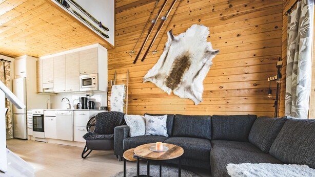 A modern living room with a large gray sectional sofa, a round coffee table, and a fur rug. A wooden ceiling with a mounted fur hide is visible.