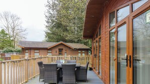 A wooden deck with a table and chairs, a glass door, and a building with a tiled roof in the background. - Oakwood Lodge (Windermere)