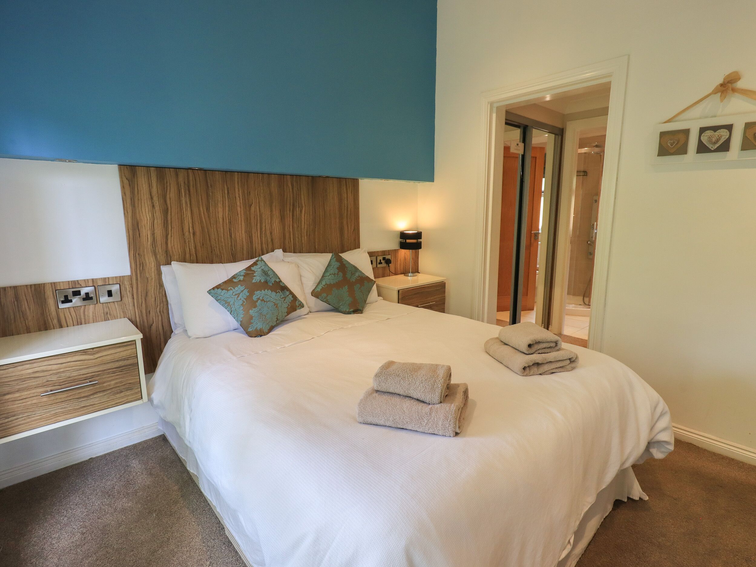 A neatly made bed with two pillows, a bedside table with a lamp, and a wall-mounted power outlet.