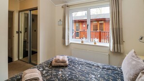 A bedroom with a bed, a window with curtains, a radiator, and a view of a wooden building outside. - Fir Tree Lodge (Windermere)