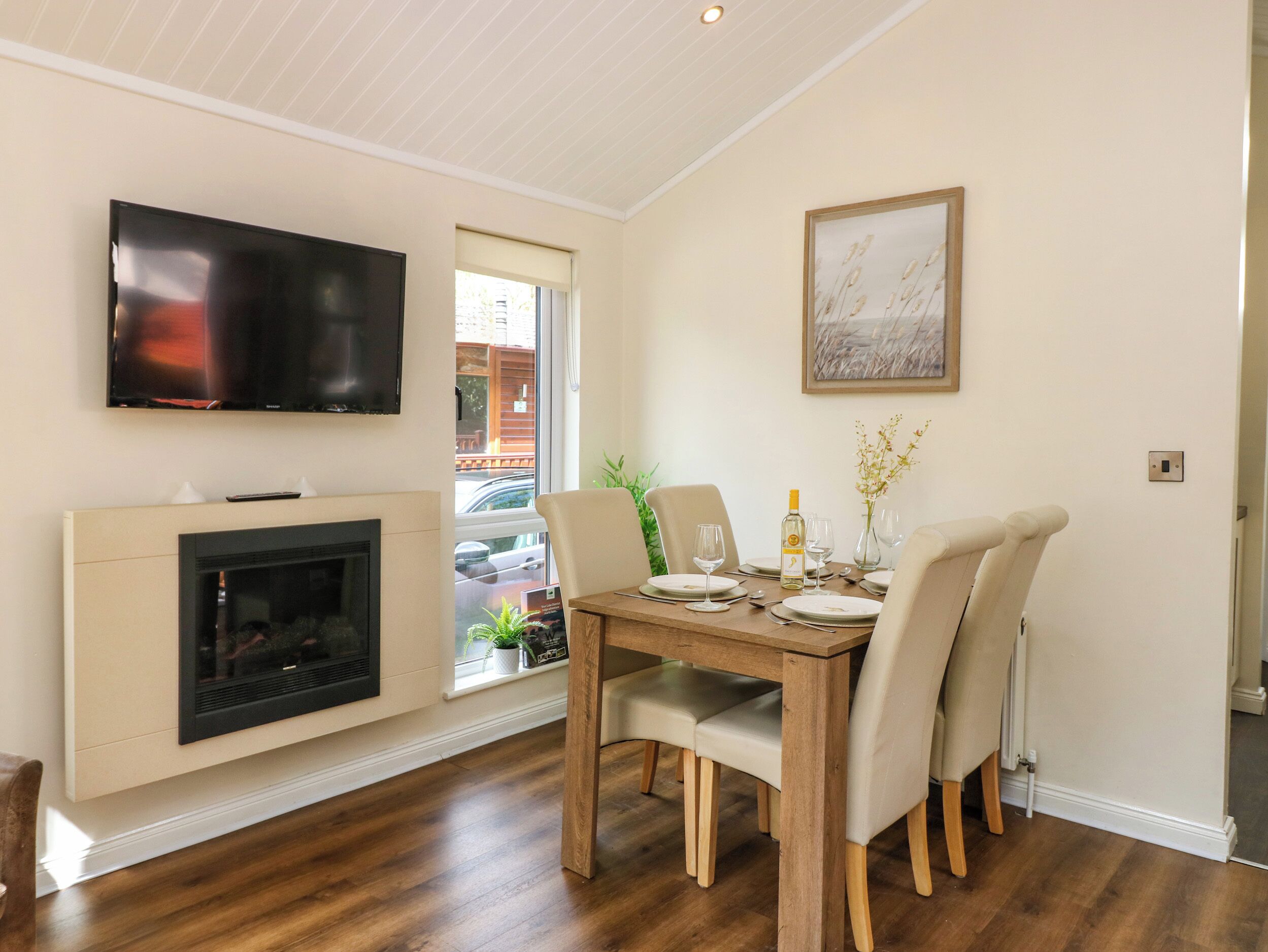 Comedor con mesa de madera dispuesta para cuatro personas, chimenea, televisor y ventana con vista a la calle.