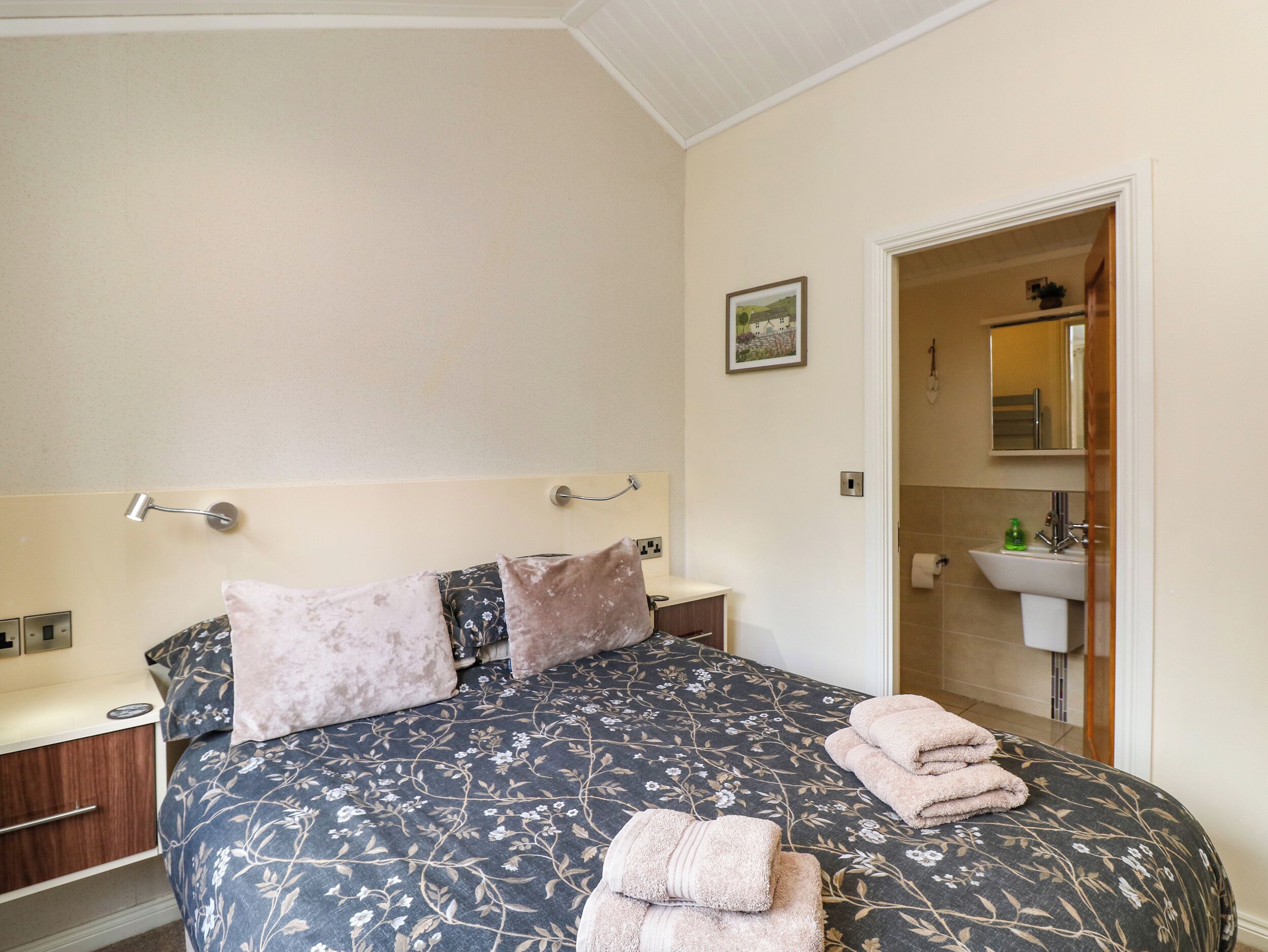 A bedroom with a bed, pillows, a bedside table, a nightstand, a bathroom with a sink and mirror, and a framed picture on the wall.