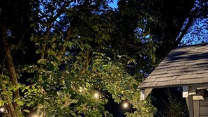 Un jardín con un sendero de grava, una pérgola de madera y luces colgantes.