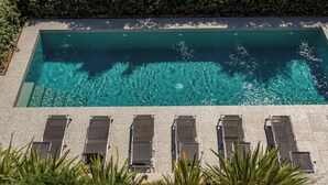 Una piscina rectangular con agua cristalina azul, rodeada por una terraza de piedra y seis sillas de descanso dispuestas en dos filas.
