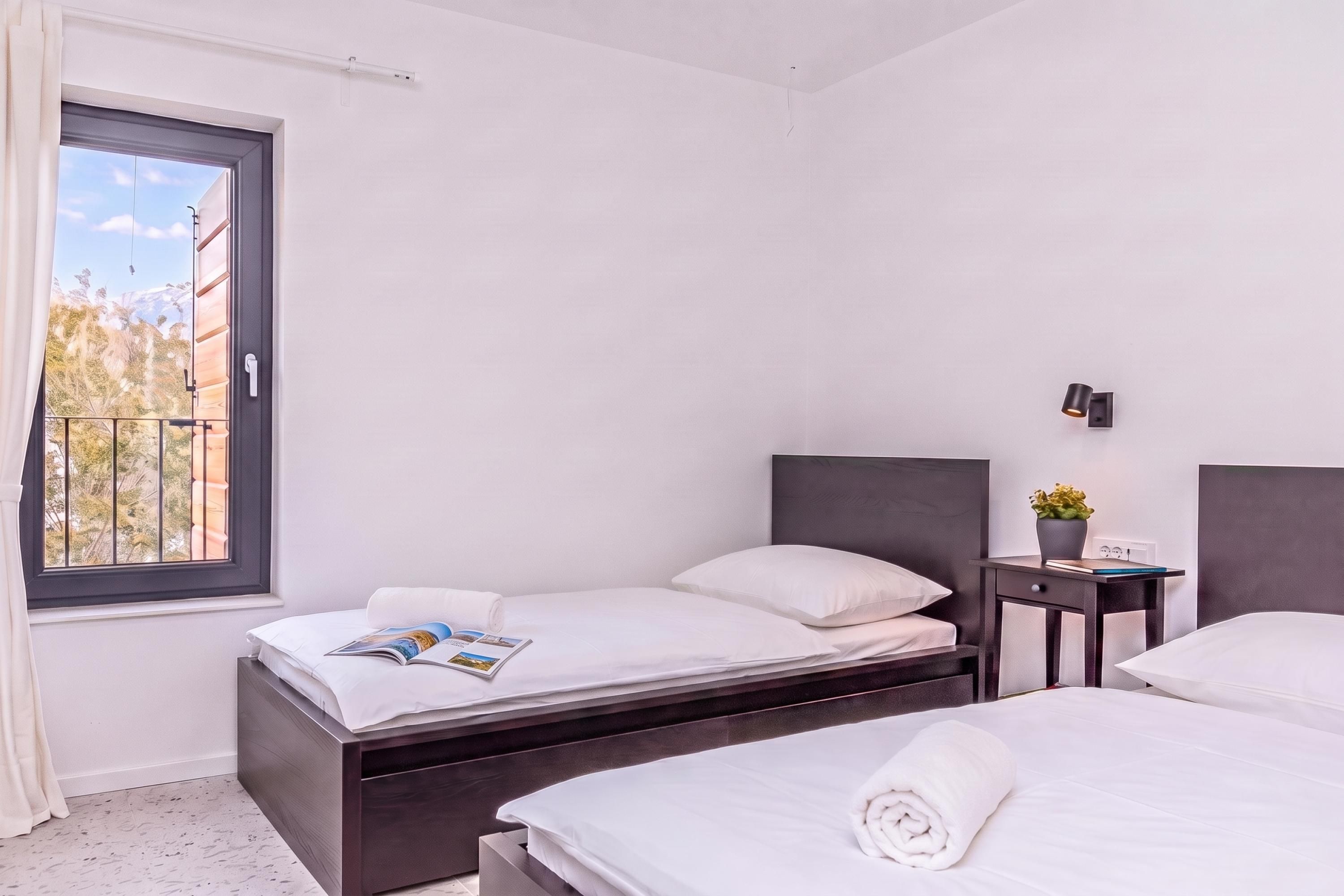 Een hotelkamer met twee bedden, een raam met uitzicht op bomen en bergen, een nachtkastje met een lamp, en een kleine plant in een pot.