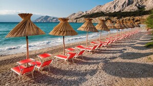 Una playa con hileras de sillas rojas y sombrillas de paja, montañas rocosas al fondo.