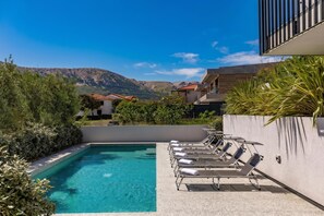 A swimming pool with lounge chairs and solar panels, surrounded by greenery and a mountain view.