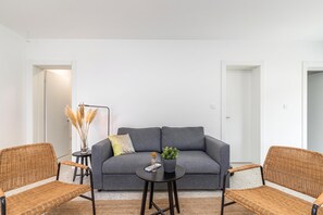 Een moderne woonkamer met een grijze bank, twee rieten stoelen, een klein rond tafeltje met een plant en een vaas met veren.
