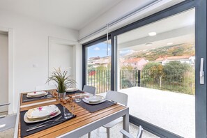 Un comedor con una mesa dispuesta para dos personas, una planta en maceta y vistas a un barrio residencial a través de grandes ventanales.